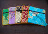 Five colorful, square satin fabric brocade pouches with floral patterns. The pouches are green, purple, black, red, and turquoise, each with a black zipper, colored tassel and gold trim.